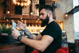 긴 수염을 가진 젊은 성인 힙스터 남자가 현대적인 카페 바에 앉아 신문을 읽고 감정적으로 반응합니다.