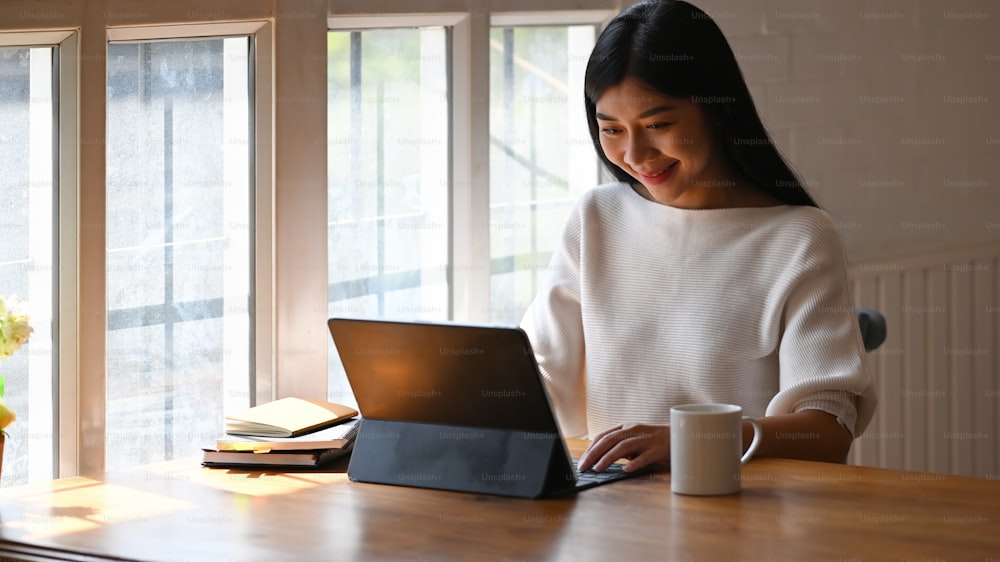 Femme créative utilisant / tapant sur une tablette d’ordinateur tout en étant assise devant une plante en pot et des livres à la table en bois moderne avec un salon confortable et des fenêtres en arrière-plan.