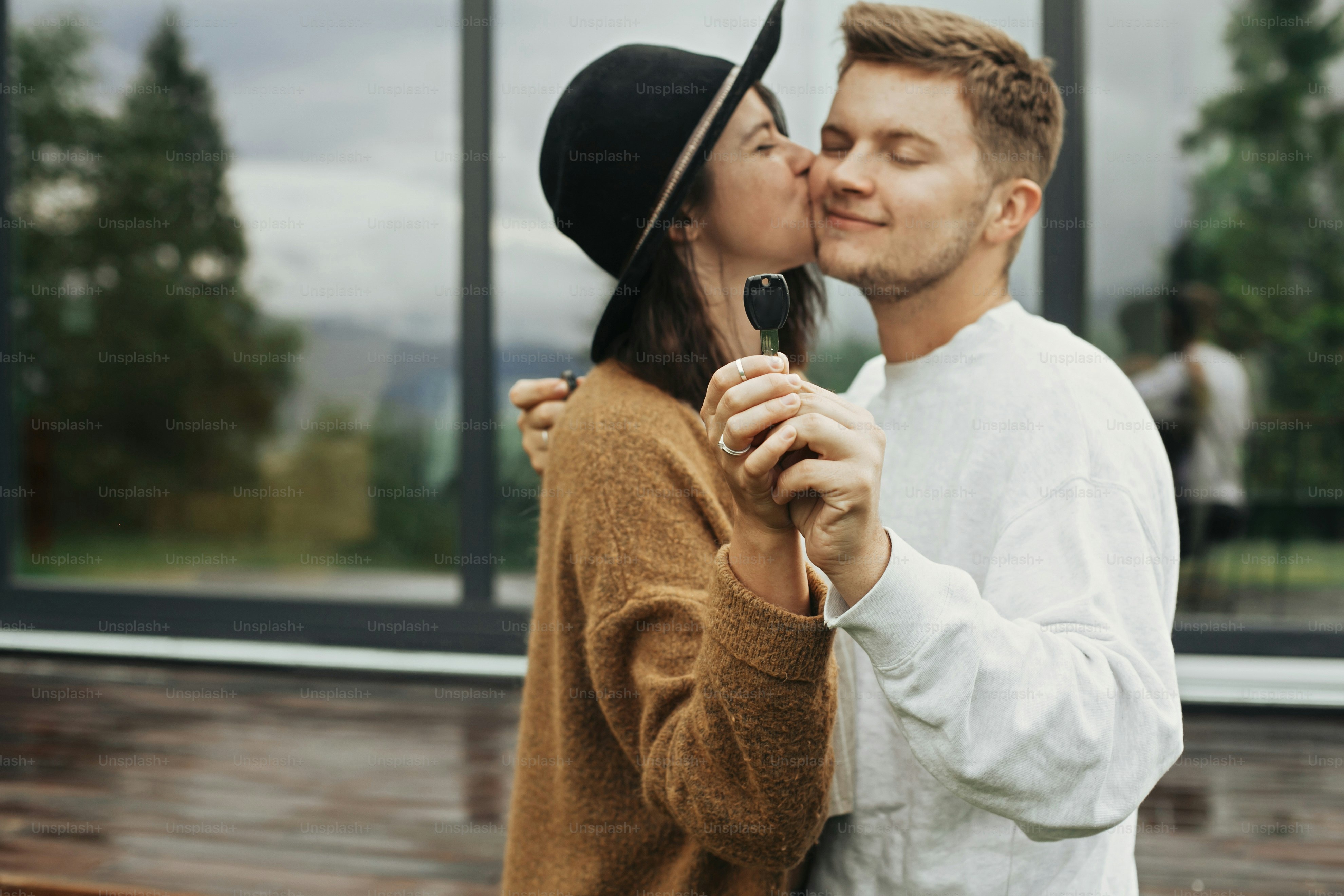 Stylish hipster couple holding together key from their new home on background of terrace and big windows outdoors. Happy young family kissing, celebrating buying first property.