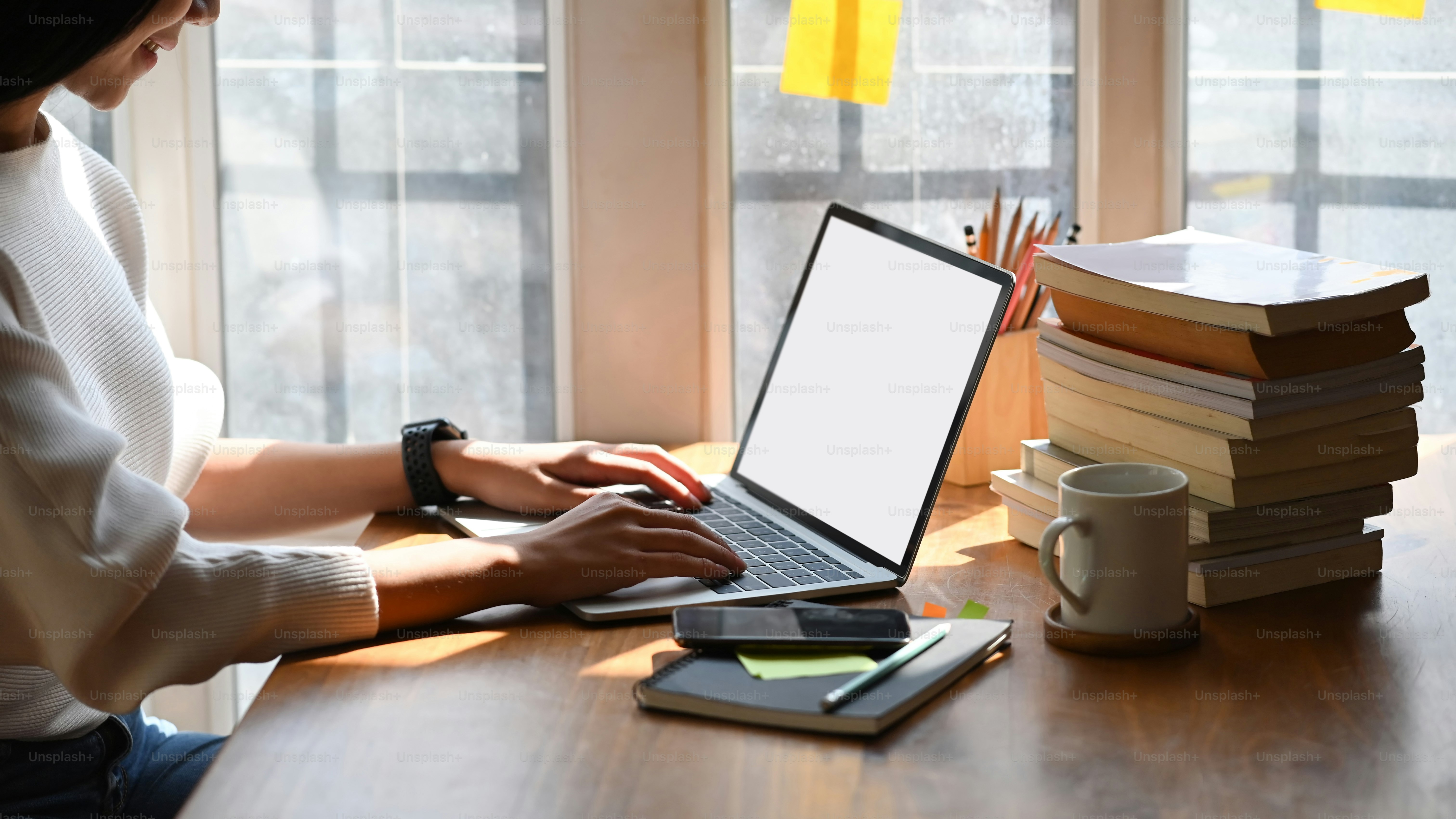 Cropped image of young beautiful woman working as writer typing on computer laptop with white blank screen while sitting at the wooden working table with sunlight through windows as background.