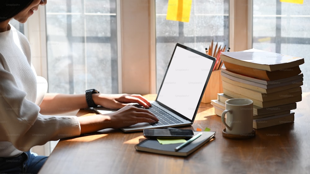 Abgeschnittenes Bild einer jungen schönen Frau, die als Schriftstellerin arbeitet, tippt auf einem Computer-Laptop mit weißem leerem Bildschirm, während sie am hölzernen Arbeitstisch mit Sonnenlicht durch Fenster als Hintergrund sitzt.
