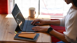 흰색 빈 화면이 있는 컴퓨터 태블릿에서 작가로 일하는 젊은 아름다운 여성의 측면 사진, 창문을 통해 햇빛을 배경으로 나무 작업 테이블에 앉아 있습니다.