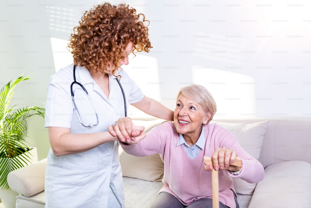 노인 여성이 거실의 소파에서 일어나도록 돕는 여성 간병인. 웃는 간호사가 노인 여성이 일어나도록 돕고 있다. 소파에서 일어나는 동안 환자를 지원하는 돌보는 간호사