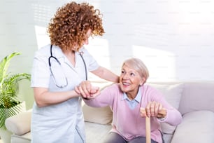 Female caregiver helping senior woman get up from couch in living room. Smiling nurse assisting senior woman to get up. Caring nurse supporting patient while getting up from sofa