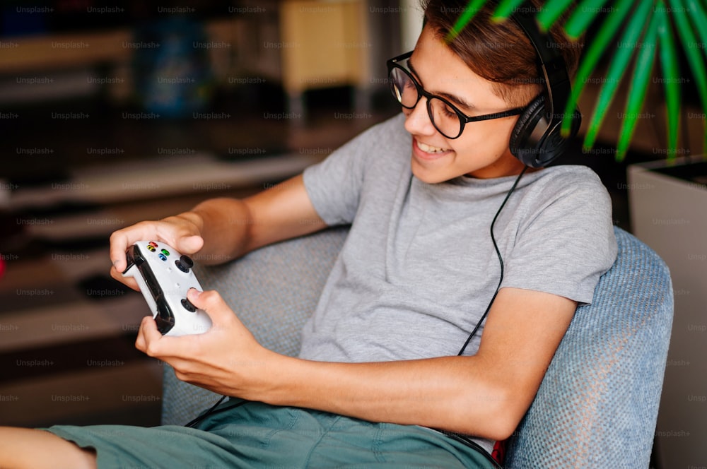 Smiling teenager boy playing console with headphones sitting in the chair indoor