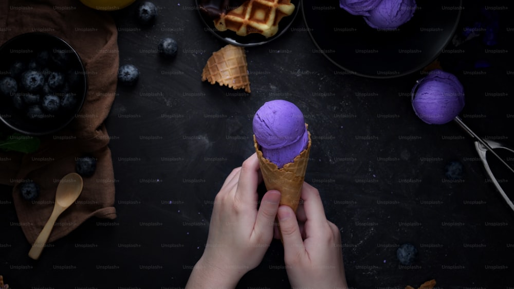 검은 테이블 배경에 블루베리, 와플, 토핑이 있는 블루베리 아이스크림 콘을 들고 있는 소녀의 상위 뷰