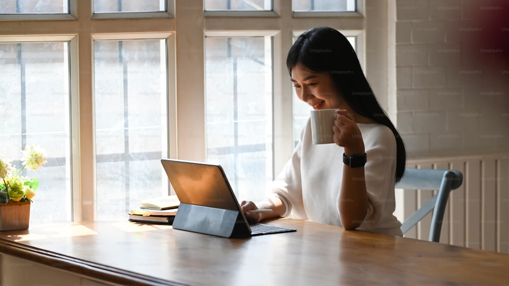 Kreative Frau in weißem Baumwollkleid, die eine Kaffeetasse in der Hand hält, während sie am Holztisch mit gemütlichem Wohnzimmer als Hintergrund arbeitet.