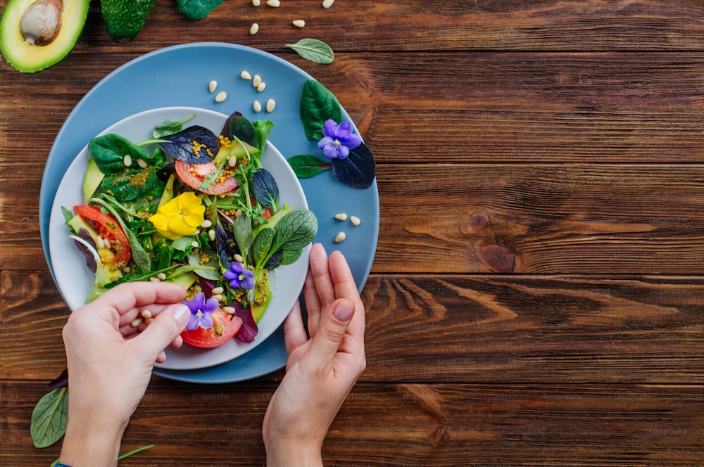 Young woman preparing fresh healthy salad with edible flowers, green, avocado, tomato and pine nuts on wooden background with blank space for text. Top view, flat lay.