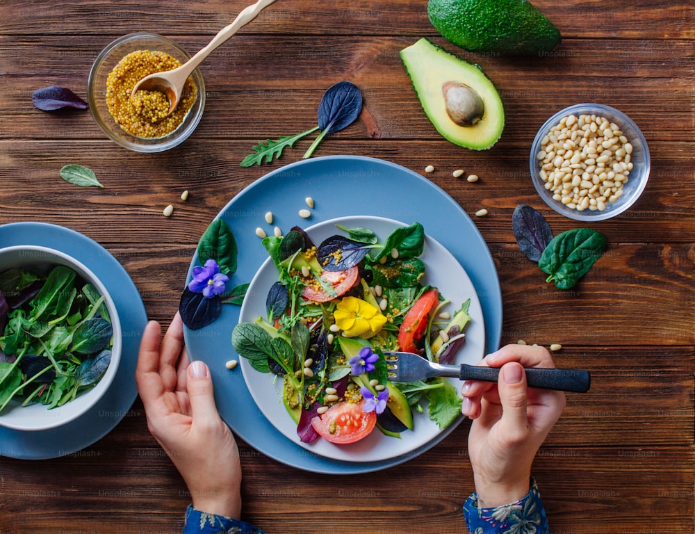 Junge Frau isst frischen gesunden Salat mit essbaren Blumen, Grün, Avocado, Tomaten und Pinienkernen auf Holzhintergrund mit leerem Platz für Text. Draufsicht, flach gelegt.