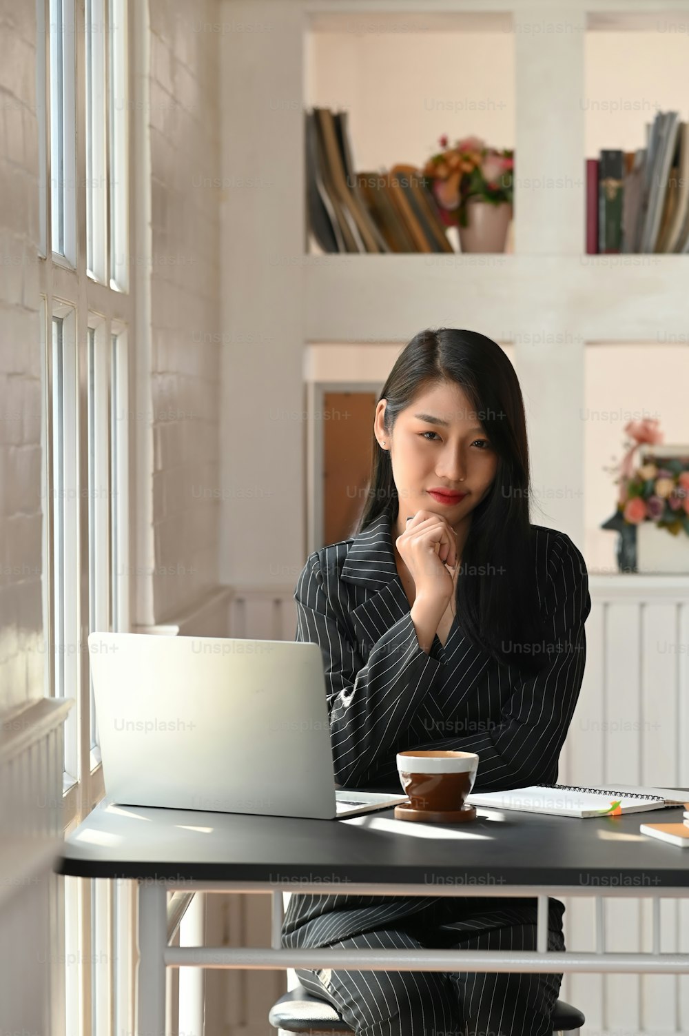 Foto de uma linda empresária sentada na mesa de trabalho moderna e mantendo a mão no queixo com sala de trabalho de luxo como fundo. Laptop, xícara de café, Documento de negócios Flat lay.
