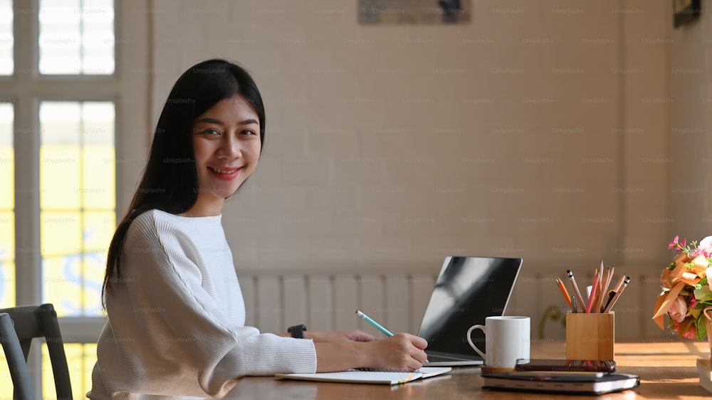 Beautiful writer sitting at the working table and writing her new fiction on notebook in front her black blank screen tablet, turning back to camera and smiling. Women lifestyle.