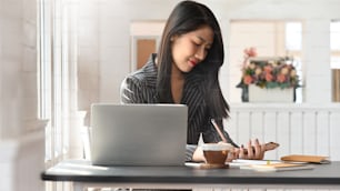 快適なオフィスを背景に、モダンな作業台でコンピューターのラップトップの前に座りながら、メモを取り、スマートフォンを手にしている若いビジネスウーマンの写真。