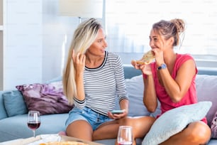 Two female friends gossiping with glasses red wine and pizzas, Showing photo images to her friend on her social media network on the mobile phone