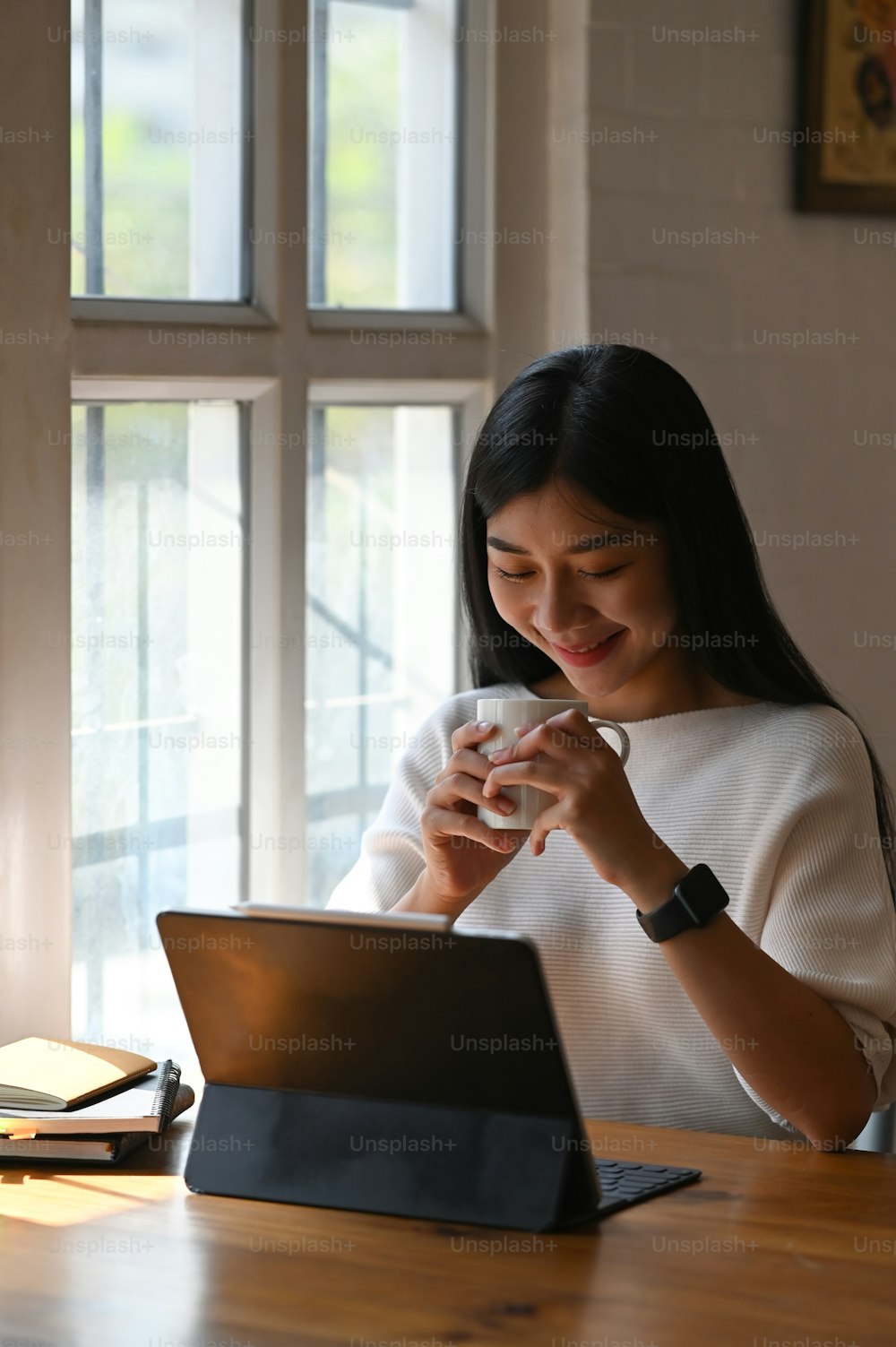 白い綿のドレスを着た若いクリエイティブな女性が、快適なリビングルームを背景に木製のテーブルで作業しながら、コーヒーカップを手にしている写真。