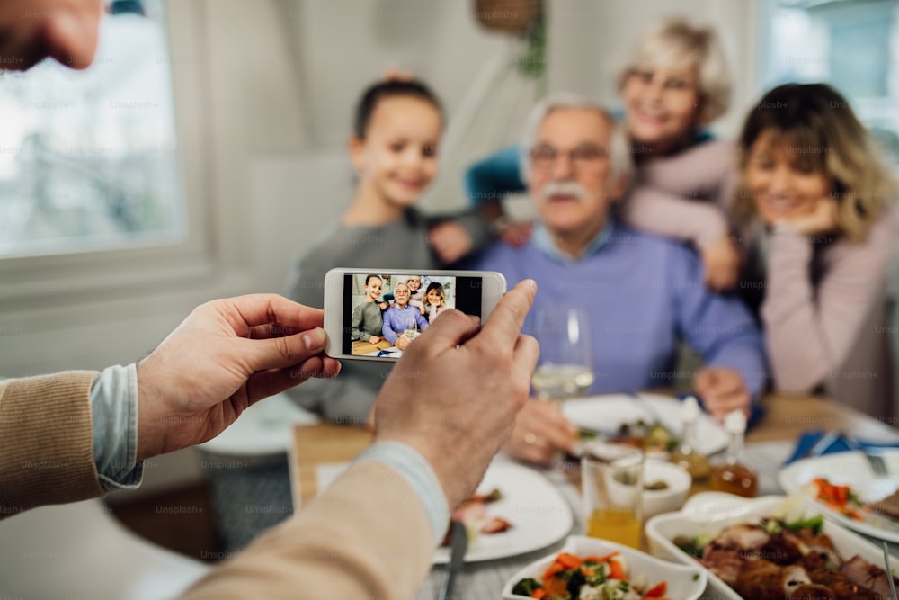 Primer plano de un hombre usando un teléfono inteligente mientras fotografía a su familia extendida en el comedor.