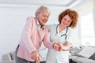 Young carer supporting senior disabled woman with walking stick. Portrait of happy female caregiver and senior woman walking together at home. Professional caregiver taking care of elderly woman.