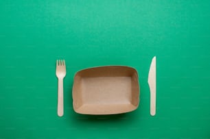Envases desechables de alimentos ecológicos. Recipiente de comida de papel kraft marrón con tenedor y cuchillo sobre fondo verde con espacio en blanco para texto. Vista superior, plano.