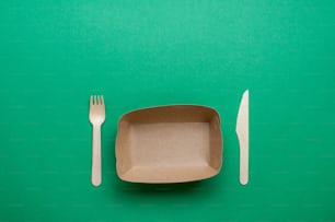 Disposable eco friendly food packaging. Brown kraft paper food container with fork and knife on green background with blank space for text. Top view, flat lay.