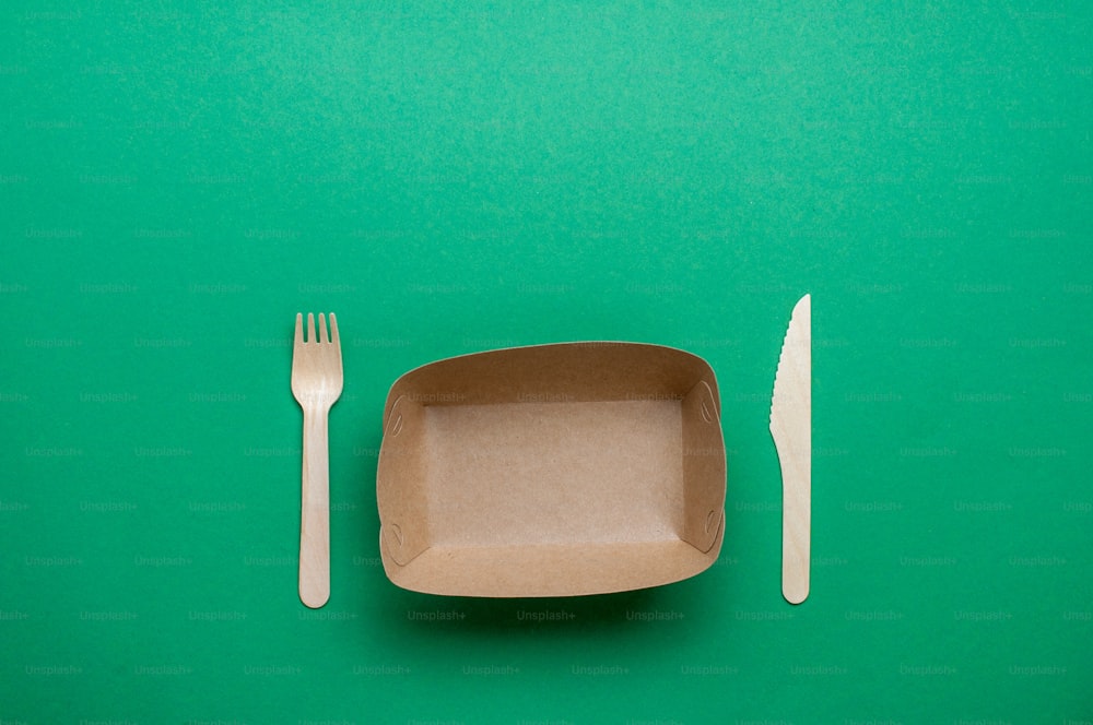 Disposable eco friendly food packaging. Brown kraft paper food container with fork and knife on green background with blank space for text. Top view, flat lay.