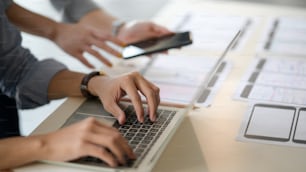 Vista de cerca de un joven desarrollador masculino de la interfaz de usuario que escribe en una computadora portátil y un documento en un escritorio blanco mientras discute con un compañero de trabajo en una sala de oficina simple