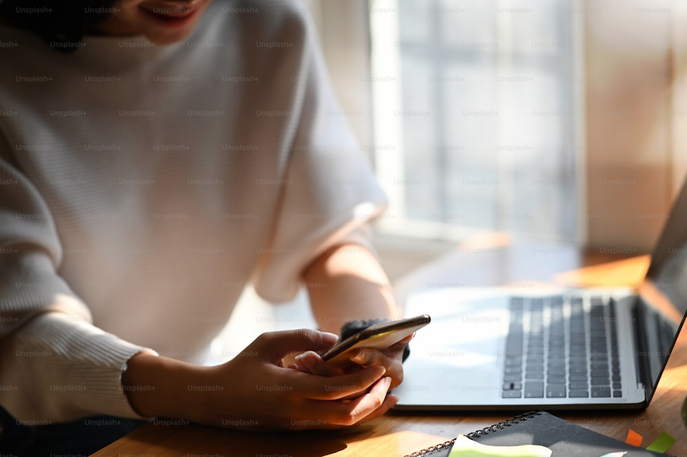 Cropped shot of young designer woman holding smartphone in hands while sitting at the modern wooden desk. Surfing the internet. Relaxing time after finished work. Break time. Woman life style.