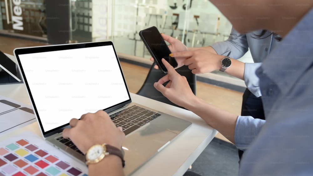 Cropped shot of UI developer working on blank screen laptop on white desk while consulting with co worker in simple office room