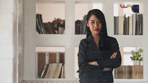 Gorgeous businesswoman in black striped suit standing with crossed arm with modern book shelf as background. Executive women, CEO, Business board concept.