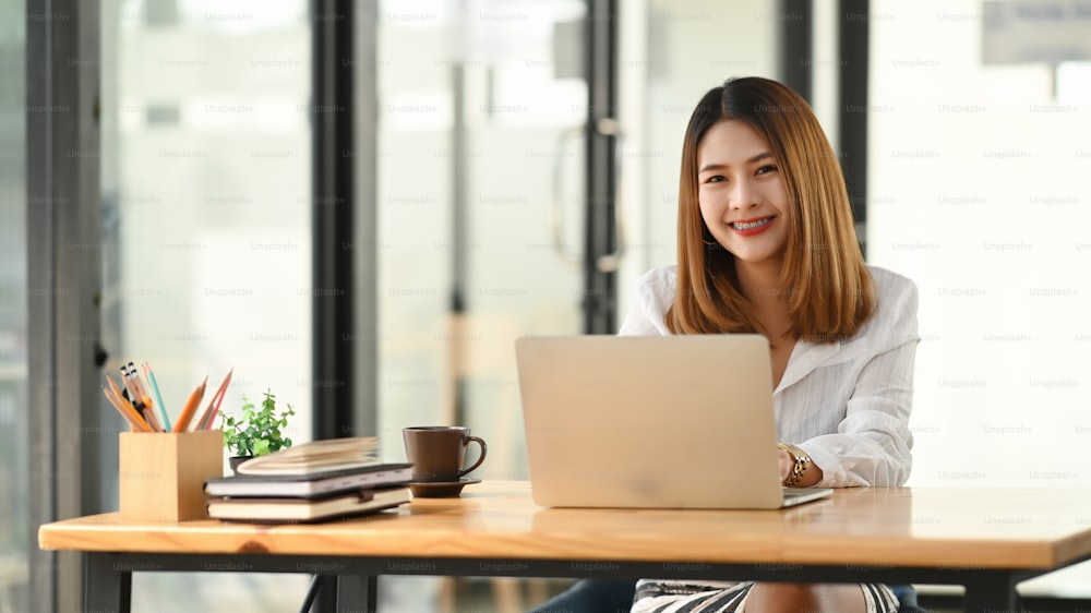 Young creative woman in white comfortable shirt working in front computer laptop at the modern wooden table with office wall glass as background.