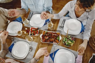 Hochwinkelansicht der Großfamilie, die Händchen hält und vor einer Mahlzeit am Esstisch betet.