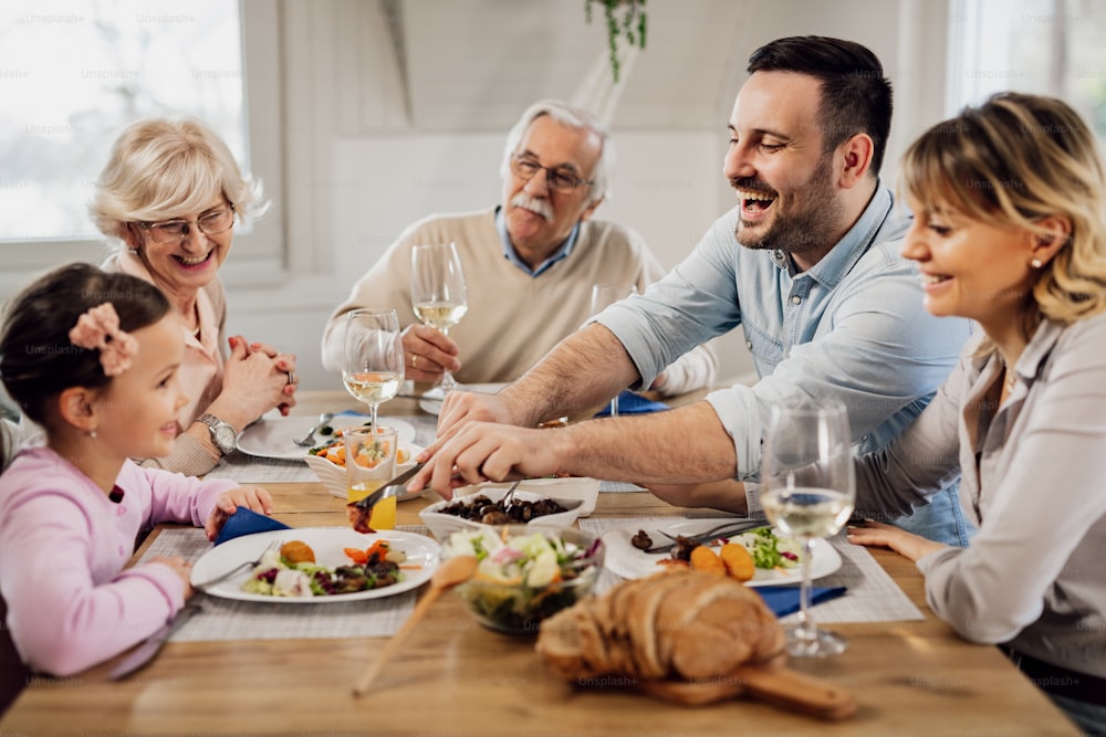 Família extensa feliz comendo juntos e se divertindo durante a hora do almoço na mesa de jantar. O foco está no homem adulto médio.