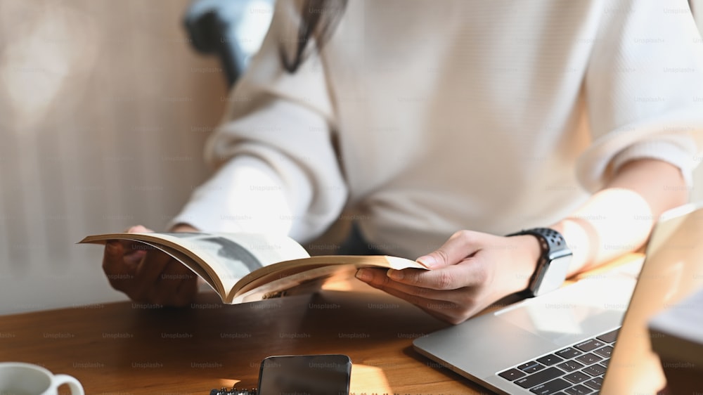 편안한 거실을 배경으로 한 현대적인 나무 테이블에서 컴퓨터 노트북, 커피 컵, 스마트폰 앞에서 손에 책을 들고 책을 읽는 젊은 아름다운 여성의 자른 이미지.