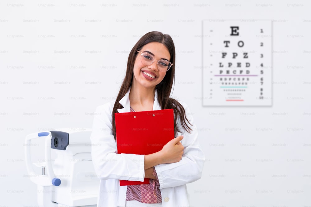 Smiling Ophthalmologist holding clipboard on the background of eye chart. Female oculist docto with an eye chart behind her