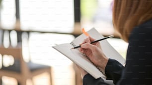 Cropped shot of female freelancer writing on mock up notebook with pencil while sitting in simple co working space
