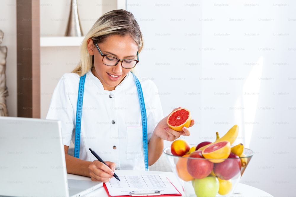 Portrait of young smiling female nutritionist in the consultation room. Nutritionist desk with healthy fruit, juice and measuring tape. Dietitian working on diet plan.