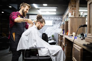 Joven barbudo que se corta el pelo por el peluquero mientras está sentado en la silla en el salón de la barbería