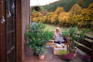 Senior woman with laptop and smartphone sitting outdoors on terrace, working.