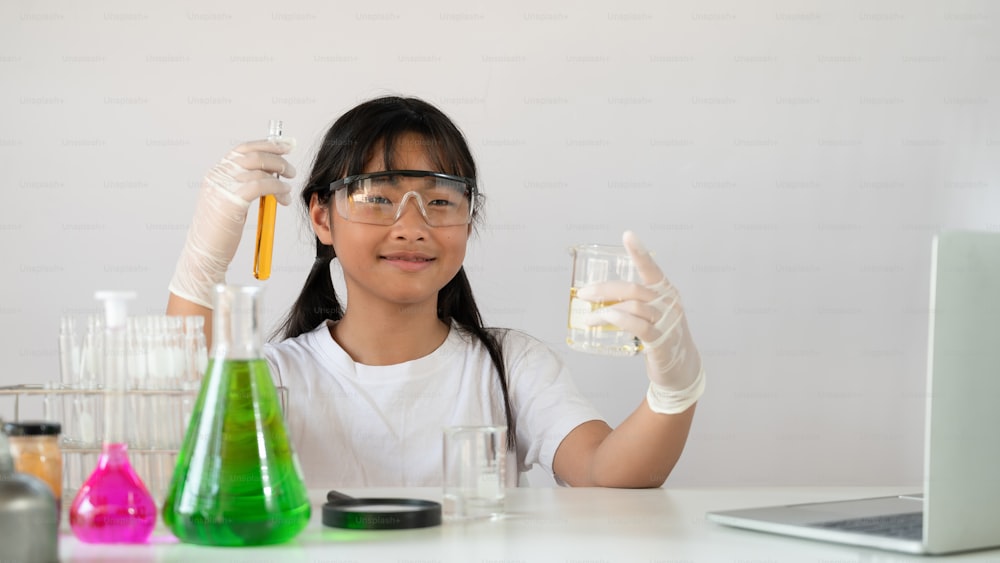 Photo of adorable schoolgirl doing a scientific experiment at the modern white table with chemistry glassware over the white laboratory wall as background. Education for kids concept.