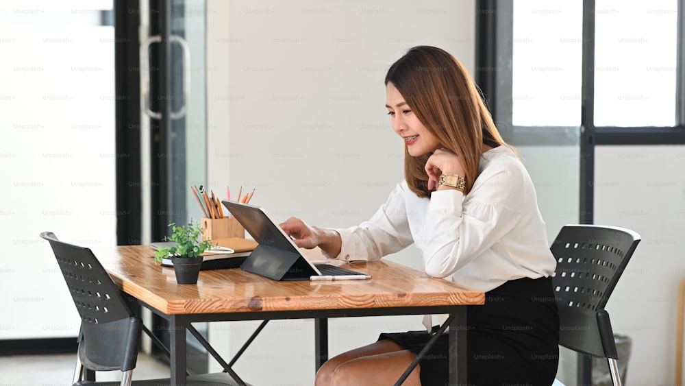 Mujer joven y hermosa que trabaja como secretaria usando una tableta de computadora con una caja de teclado mientras está sentada en la mesa de trabajo de madera con una oficina ordenada como fondo.