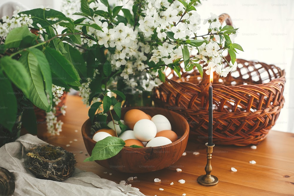 桜の花と緑の葉が付いた素朴なテーブルの上に、天然のイースターエッグ、ヴィンテージキャンドル、籐のバスケット。ハッピーイースター、雰囲気のある瞬間。田舎の静物画。白と茶色の卵