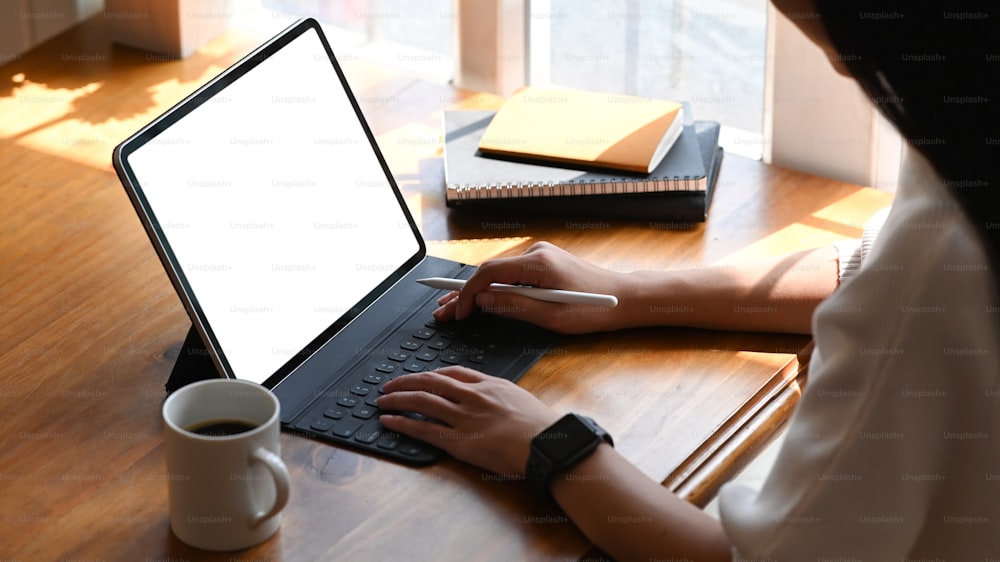 Abgeschnittenes Bild einer jungen schönen Frau, die als Schriftstellerin arbeitet und auf einem Computer-Tablet mit weißem leerem Bildschirm tippt, während sie am hölzernen Arbeitstisch mit Sonnenlicht durch Fenster als Hintergrund sitzt.