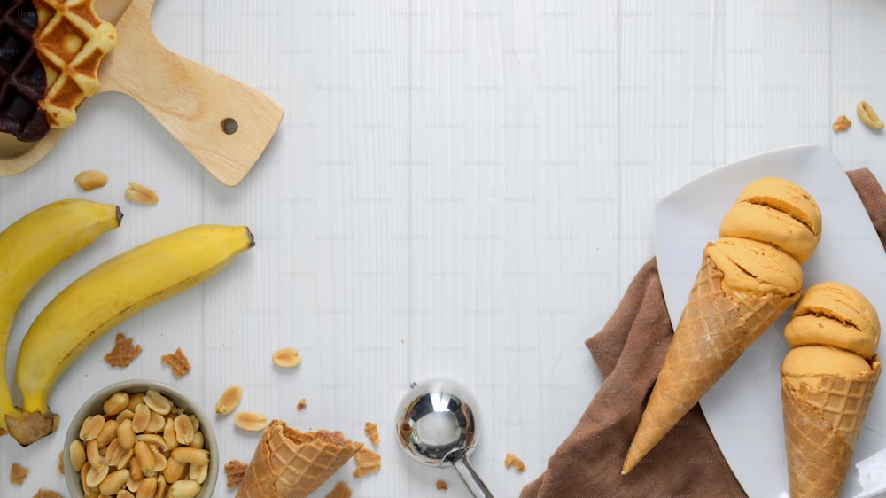 Overhead shot of summer dessert with peanut butter banana flavour ice-cream cones, waffle, topping and copy space on plank desk background