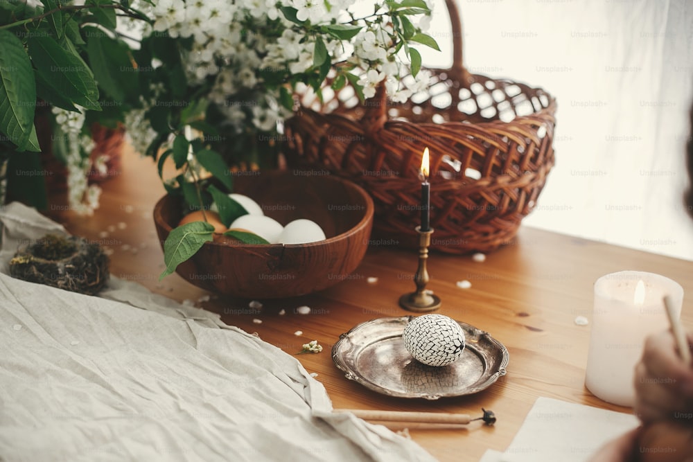 Easter Stylish Rural still life. Stylish Easter egg with modern wax ornaments and natural dyed eggs on rustic wooden table with spring flowers, basket, candle, linen cloth.  Zero waste