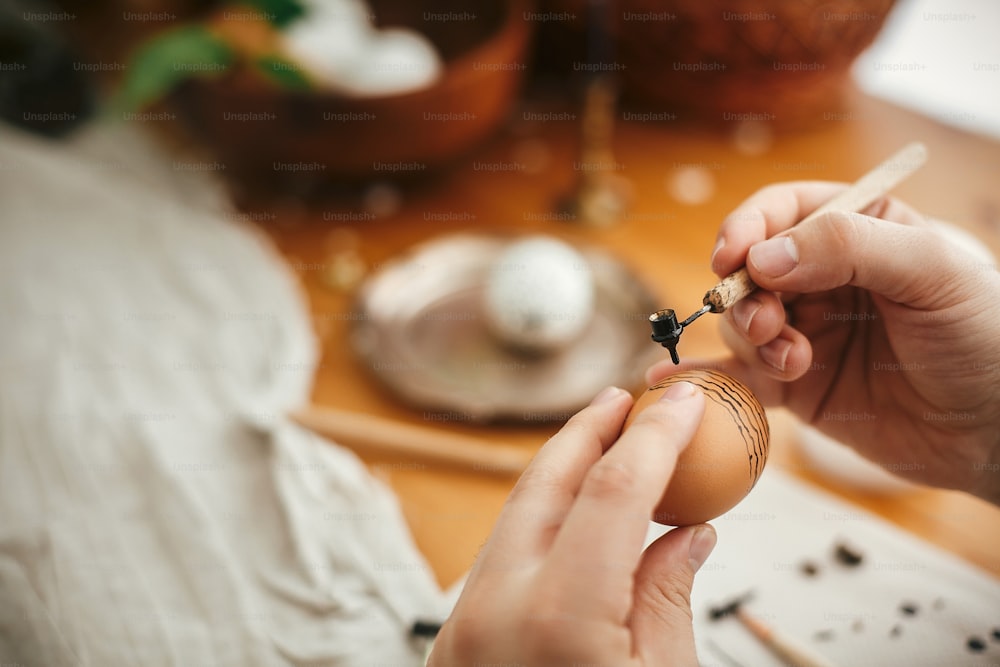 촛불, 바구니, 녹지가 있는 소박한 나무 테이블의 배경에 뜨거운 왁스로 부활절 달걀을 칠합니다. 손에 현대 장식과 부활절 달걀입니다. 행복한 부활절. 우크라이나 전통 pysanka. 폐기물 제로