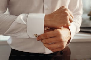 stylish groom getting ready in morning, putting on white shirt golden cufflinks. morning preparation for wedding ceremony in hotel room.