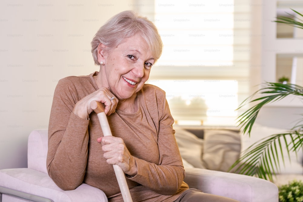 Portrait of beautiful senior woman with white hair and walking stick. Portrait of senior woman sitting on sofa at home. Smiling middle aged mature grey haired woman looking at camera