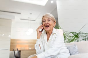 Senior woman talking on her mobile phone. Senior woman has a happy conversation at cellphone. Smiling senior woman using phone sitting on couch at home.