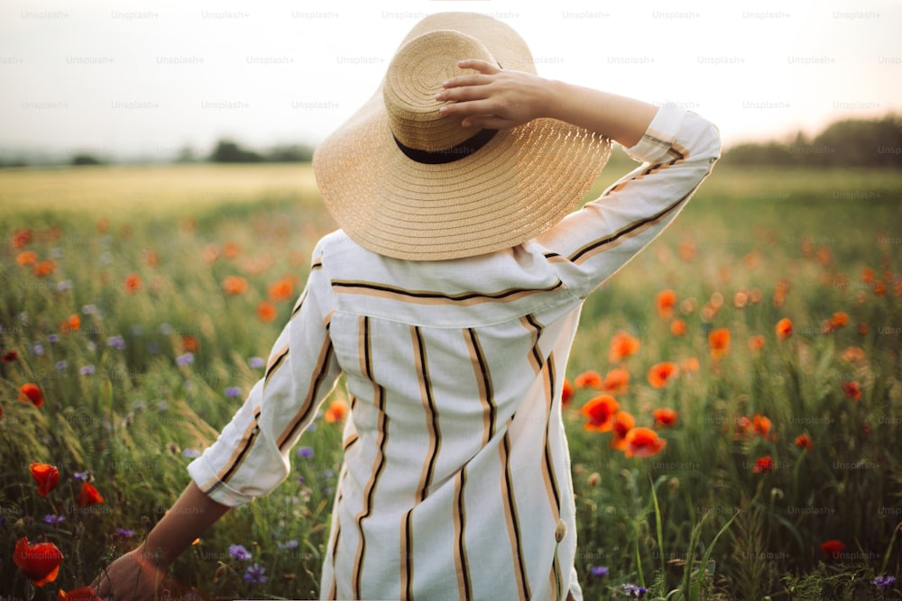 Mulher nova no vestido de linho e chapéu desfrutando da noite rural entre papoula e cornflowers no campo. Menina elegante no vestido rústico andando em flores silvestres na luz quente no prado de verão.