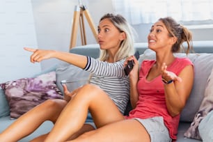 Portrait of funny and happy young women watching comedy in bed and laughing. Cheerful friends eating tasty popcorn and looking movie with gladness. Cozy and friendly atmosphere