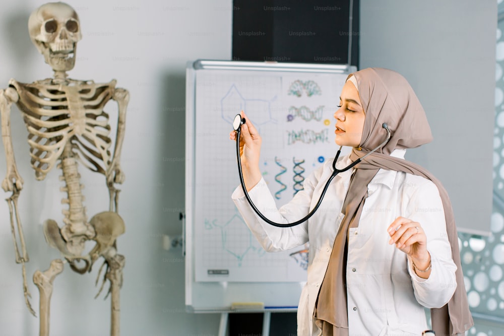 Belle jeune femme musulmane du Moyen-Orient, étudiante en médecine ou médecin, posant à l’hôpital ou dans une salle de classe avec un stéthoscope. Squelette humain et tableau de classe en arrière-plan.
