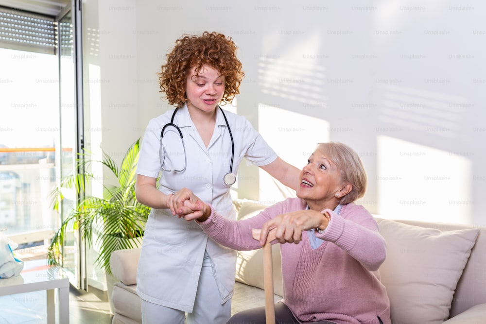 노인 여성이 거실의 소파에서 일어나도록 돕는 여성 간병인. 웃는 간호사가 노인 여성이 일어나도록 돕고 있다. 소파에서 일어나는 동안 환자를 지원하는 돌보는 간호사